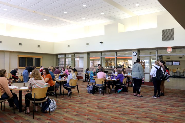 A group of students is seated at various tables in a spacious, brightly lit café with a high ceiling. Some are engaged in conversation while others focus on their meals or studies. In the background, a few students stand near the entrance, chatting. The overall atmosphere is casual and social.